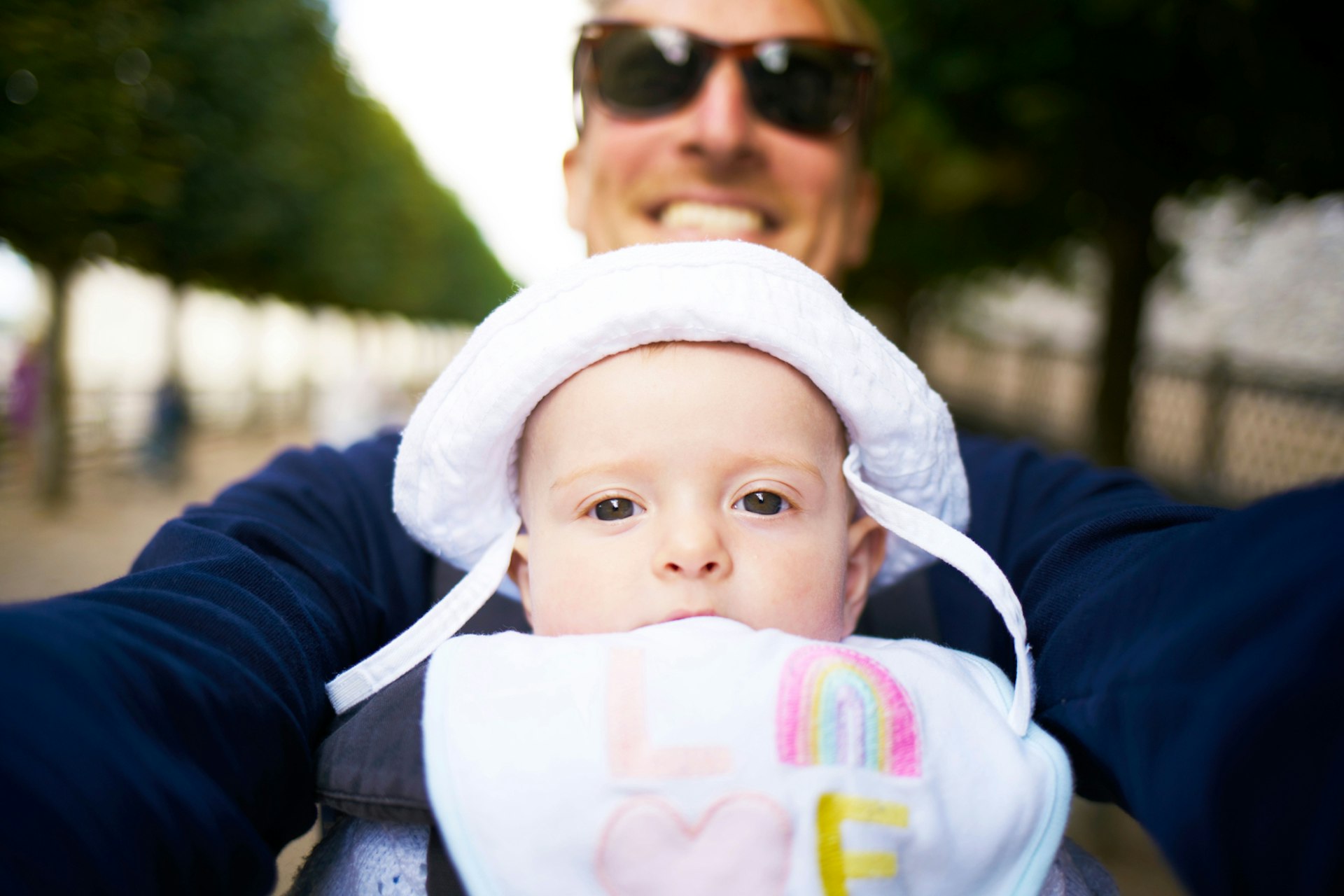 A smiling dad with sunglasses who is slightly blurred stands behind his bewildered looking baby who is looking at the camera from a sling on his chest