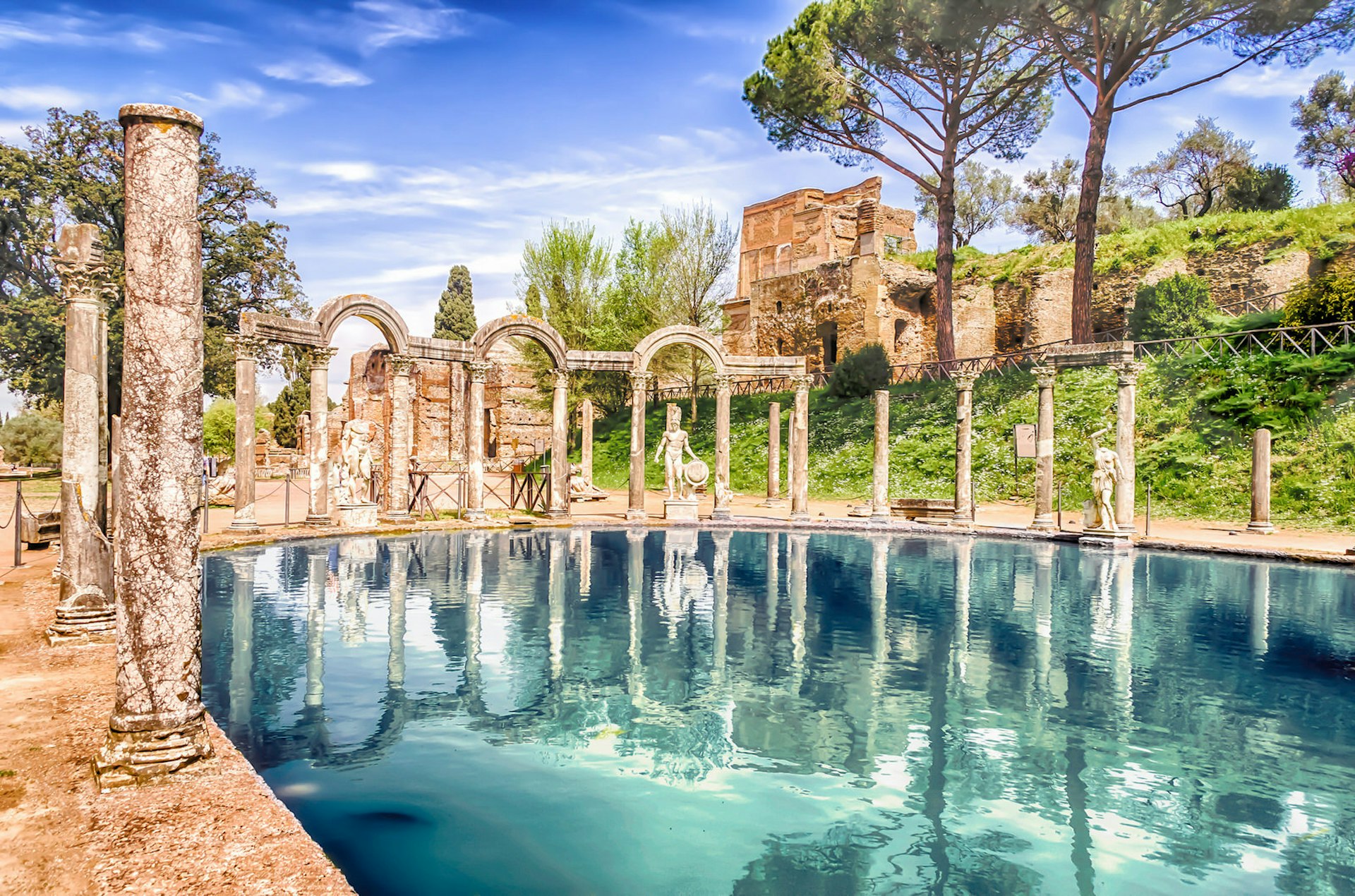 A shimmering blue pool at the Villa Adriana in Tivoli, surrounded by Roman columns, walls and statuary, with trees and grassy slopes in the background; it is the location for the Killing Eve season two finale.