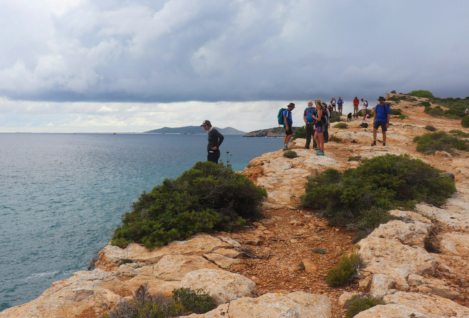 Ibiza spa breaks - a group of people walk along a cliff in Ibiza. The cliff has warm dusty orange rocks with hardy bushes and overlooks the sea and a cloudy sky