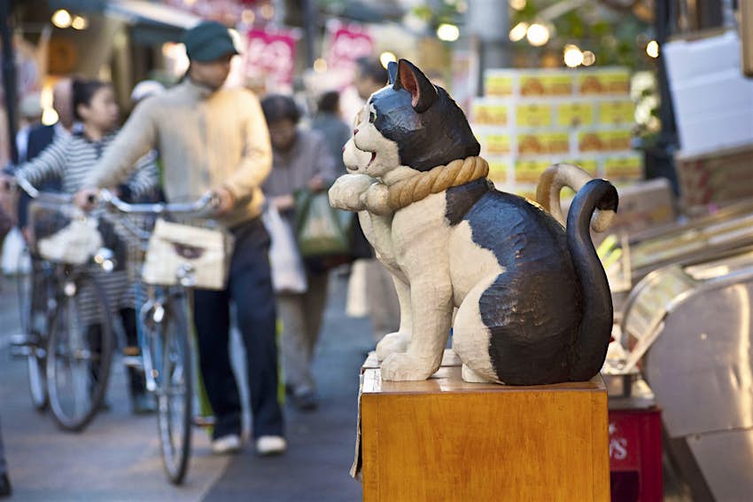 A small statue of a cat stands in front of a stall along Yanaka Ginza market street, with a group of shoppers with bicycles blurred in the background