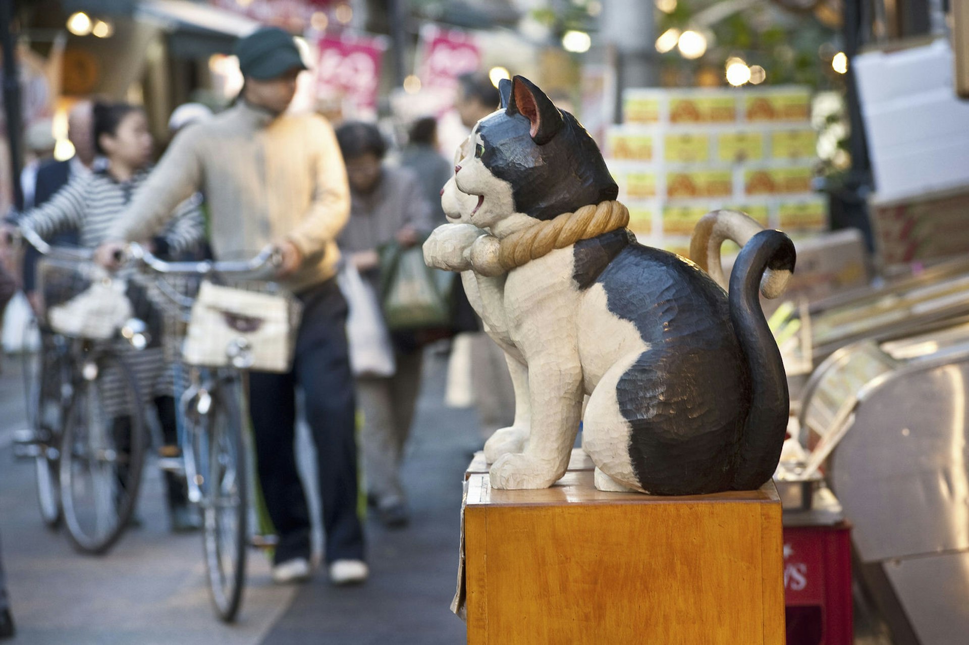 A small statue of a cat stands in front of a stall along Yanaka Ginza market street, with a group of shoppers with bicycles blurred in the background