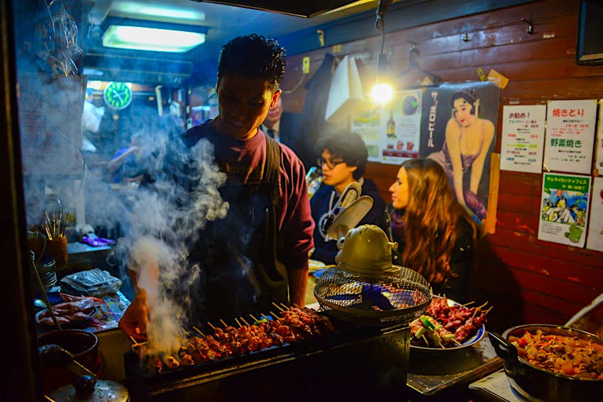 A Japanese man grills BBQ meat for customers waiting in the background at a small food stall at Omoide Yokocho