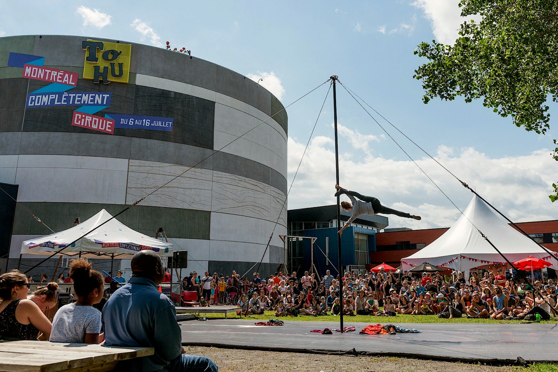 A circus acrobat performs on a vertical pole surrounded by tents and crowds, in front of the circular TOHU building in Montreal
