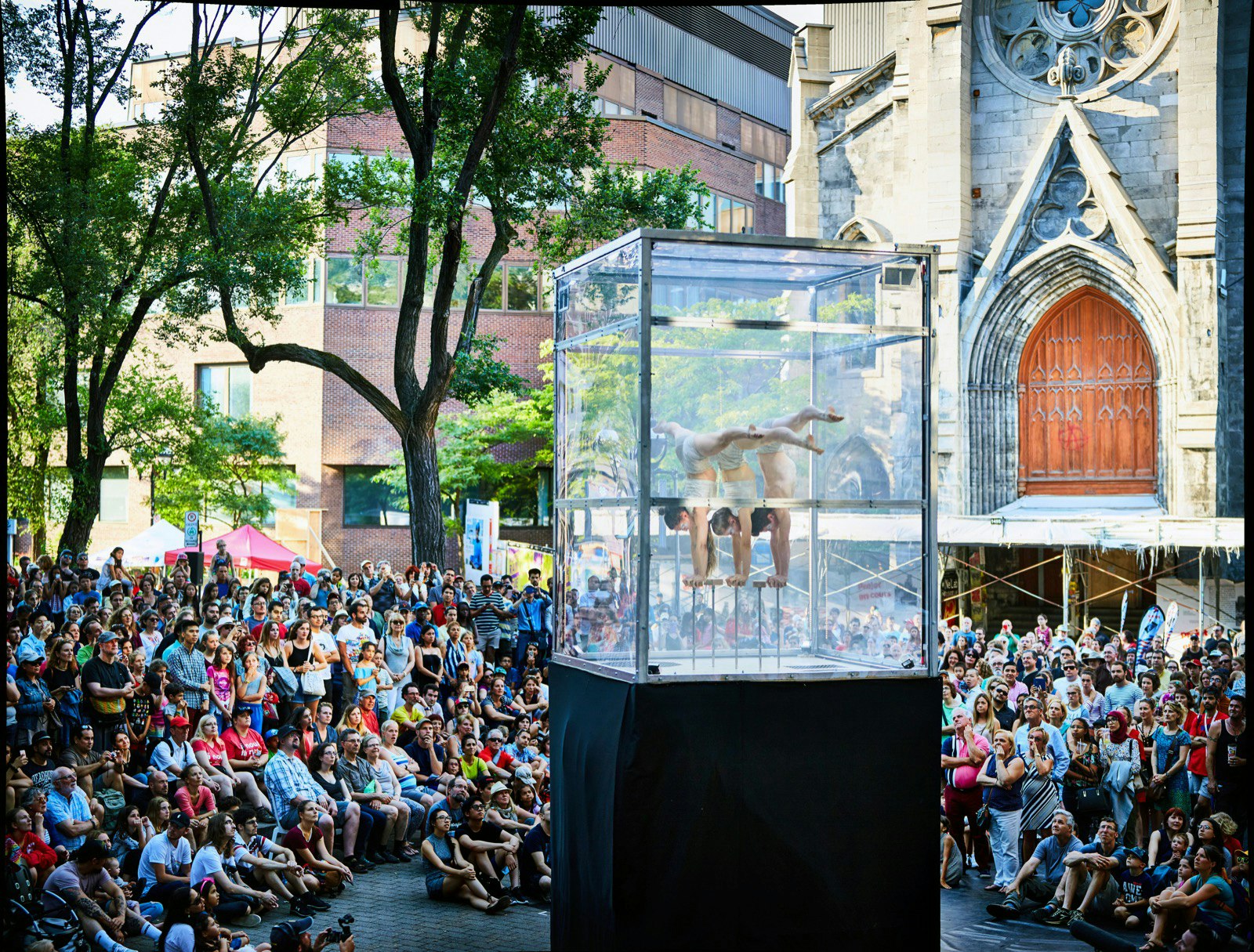 Three circus acrobat/contortionists balance on their hands on small stands inside a clear box on a pedestal, surrounded by spectators in Montreal
