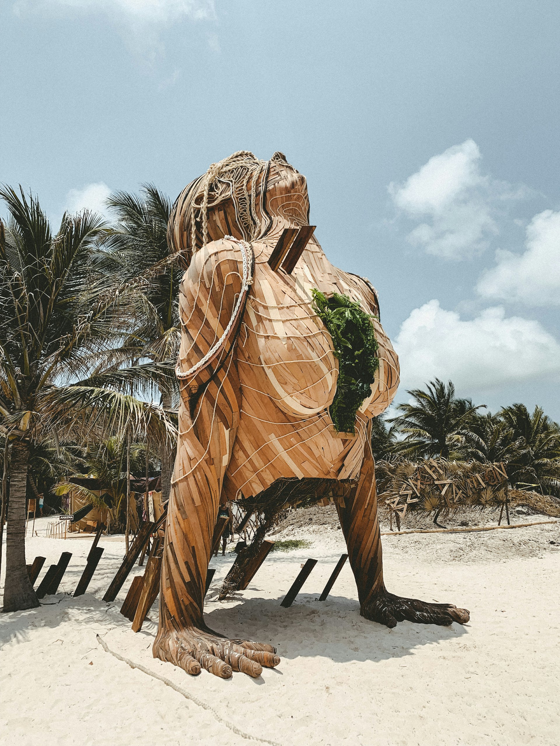 A huge wooden statue on a sunny beach of a woman's torso with hands splayed out on the sand, and face upturned to the sky; it wears a necklace of green leaves, and there are palm trees behind.