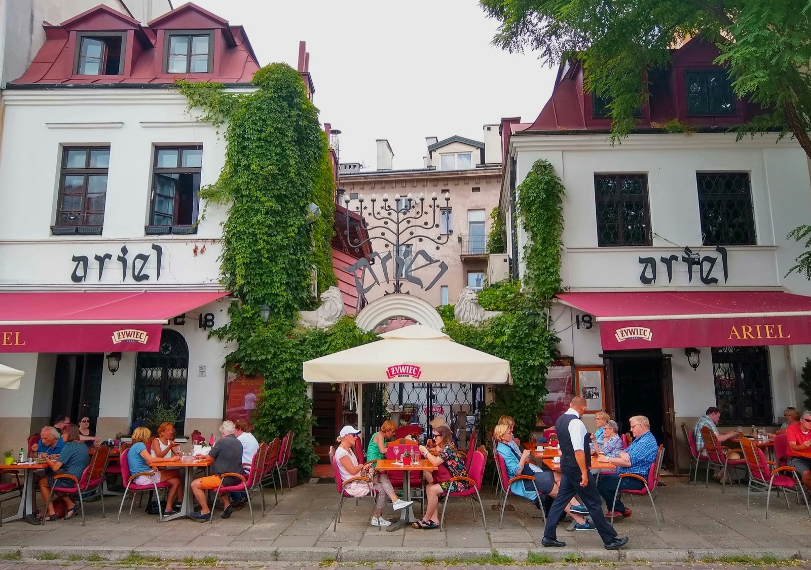 A menorah marks the top of a fence between two restaurant buildings, both with red awnings and signs that identify it as Ariel; Krakow Jewish Cultural revival