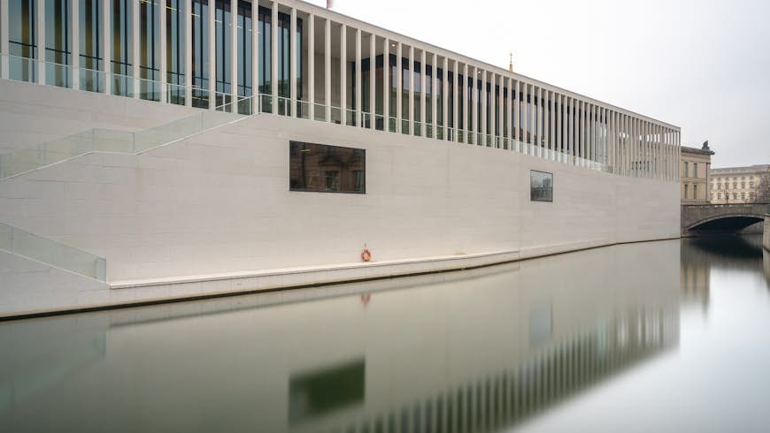 A photo of Museum Island's newest museum, the James-Simon-Galerie, taken from across the water. The building is very modern with large cream-beige bricks creating sleek lines. The windows are obscured by evenly spaced pillars/.