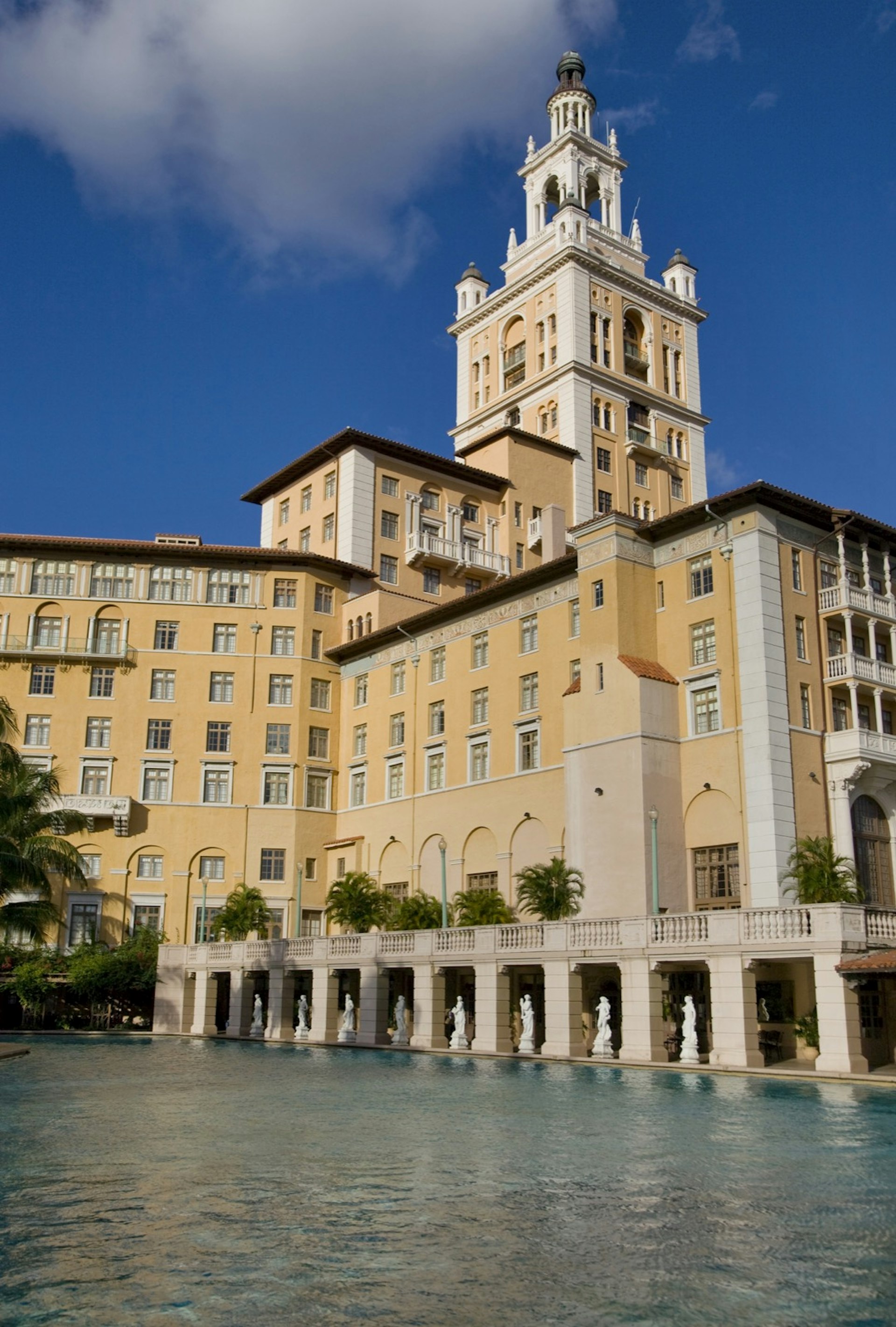 A very large pool runs right up to the edge of a cream colored building with a tall tower, as statues stand along the pool's edge. Weekend in Miami