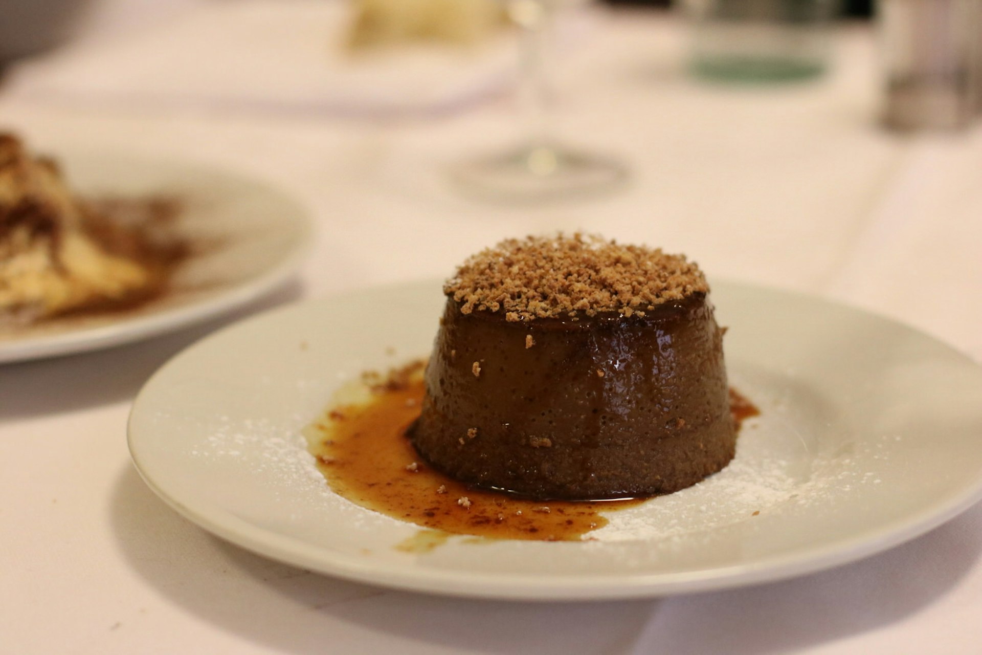 A close up shot of an Italian dessert - Bonet, served at Porto di Savona restaurant in Turin. It is a small brown cake on a white plate topped with crumbled nuts and sauce. 