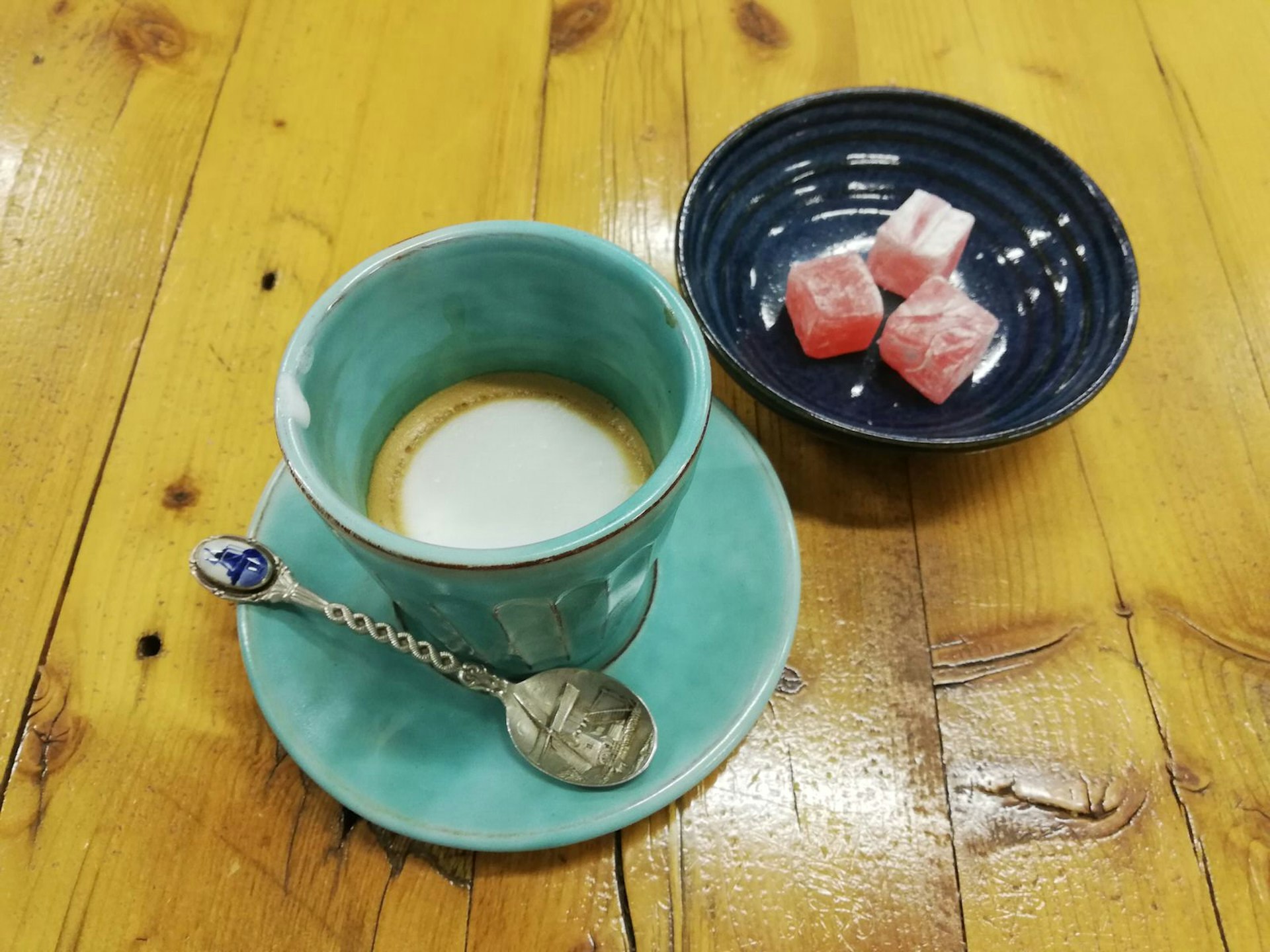 A short coffee with a small topping of milk sits in a jade-coloured cup on a matcher saucer with spoon; next on the wooden table is a deep blue bowl with some pink Turkish delight; an example of cultural variety in the Cape Town coffee scene