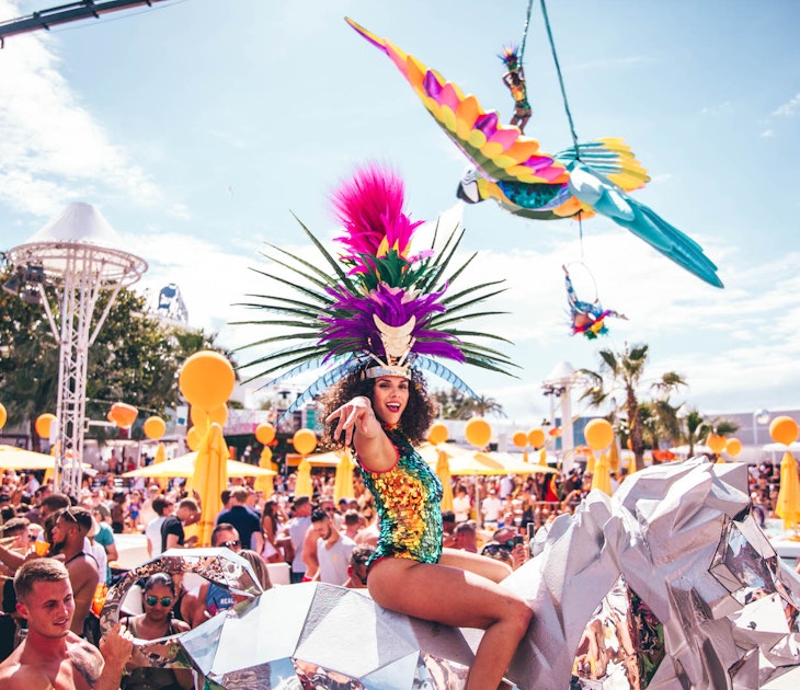A woman in a colourful sequined leotard and a huge multicoloured feather headdress sits on top of a silver unicorn in front of a huge crowd at a pool party in O Beach Ibiza during the day