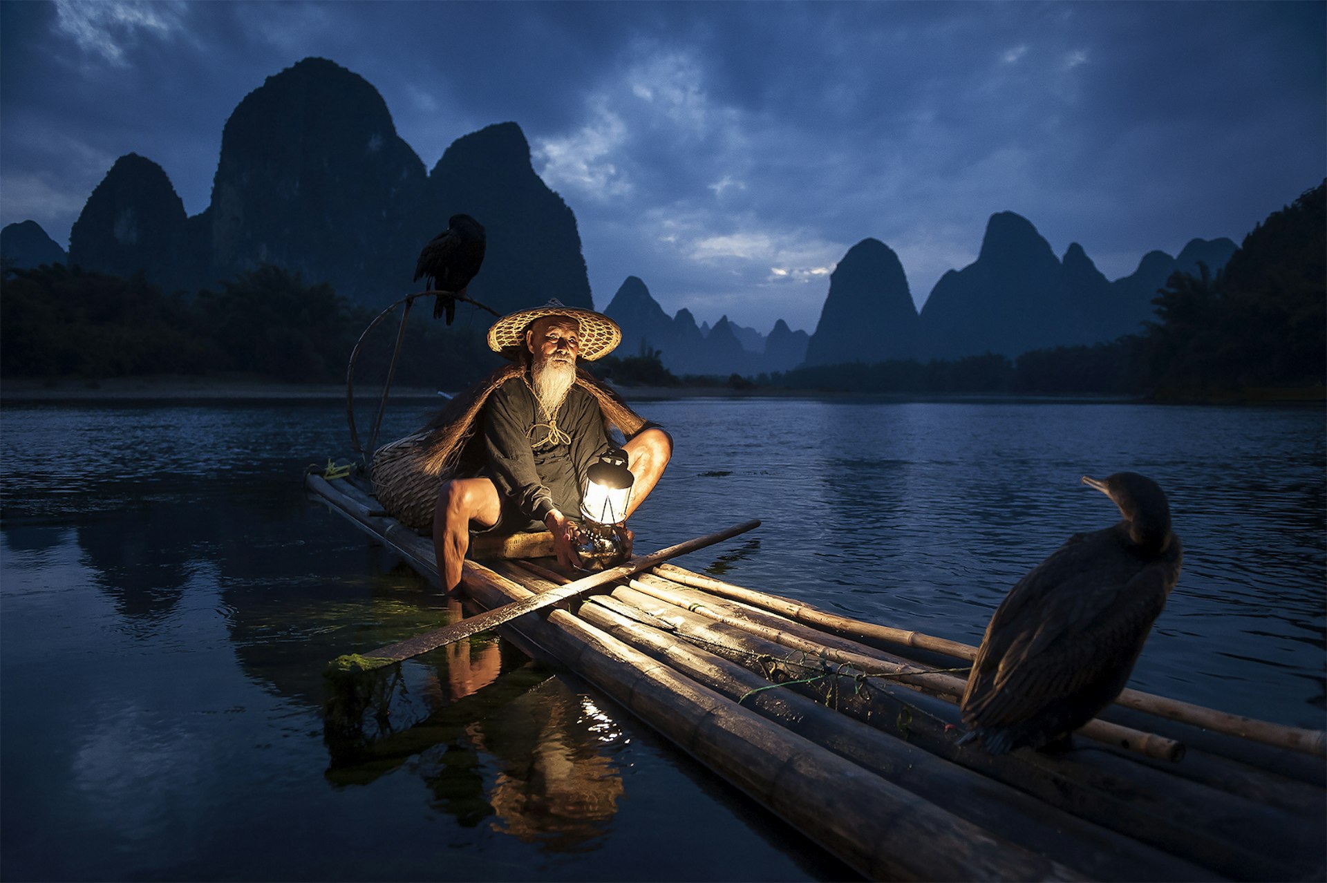 A boatman in Guilin, China; he sits in his long traditional boat made from tree branches at night, with a lantern illuminating him; he wears a wide-brimmed hat. There is a bird of prey sat at oneend of the vessel while in the background are a stretch of water and dramatic karst mountains.