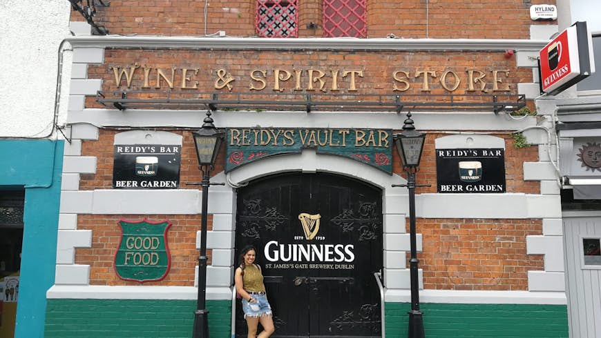 Christina Webb stands in front of Reidy's Bar; it's housed in an old brick building with a sign saying Wine & Spirit Store; there is white and green paintwork and black doors bearing the Guinness harp logo. 