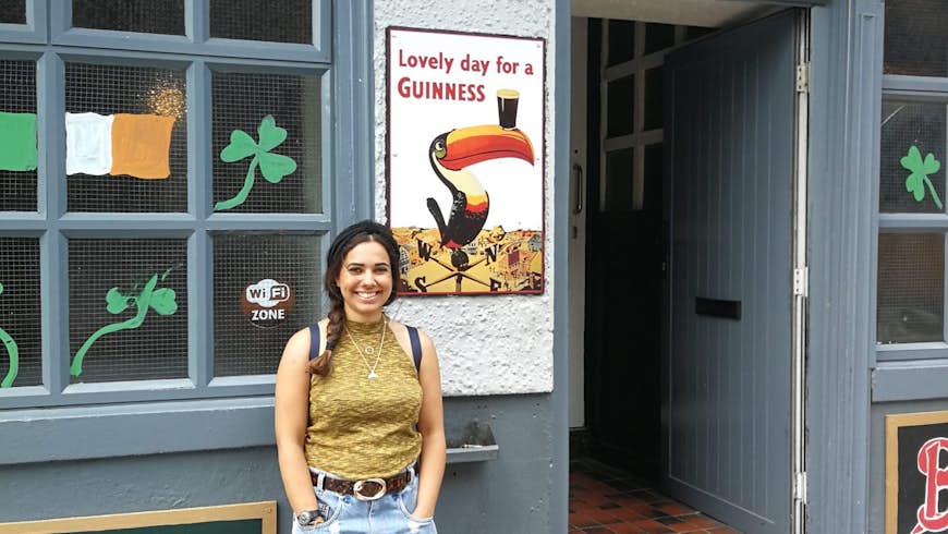 Christina stands outside the Shandon Arms; it's a building painted grey with flowers behind a fence above the door, and an Irish flag and shamrocks in the window. 