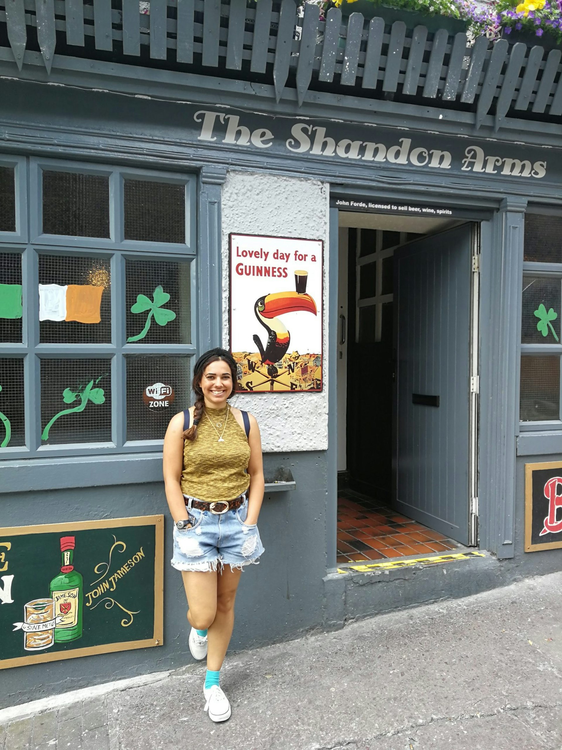 Christina stands outside the Shandon Arms; it's a building painted grey with flowers behind a fence above the door, and an Irish flag and shamrocks in the window. 