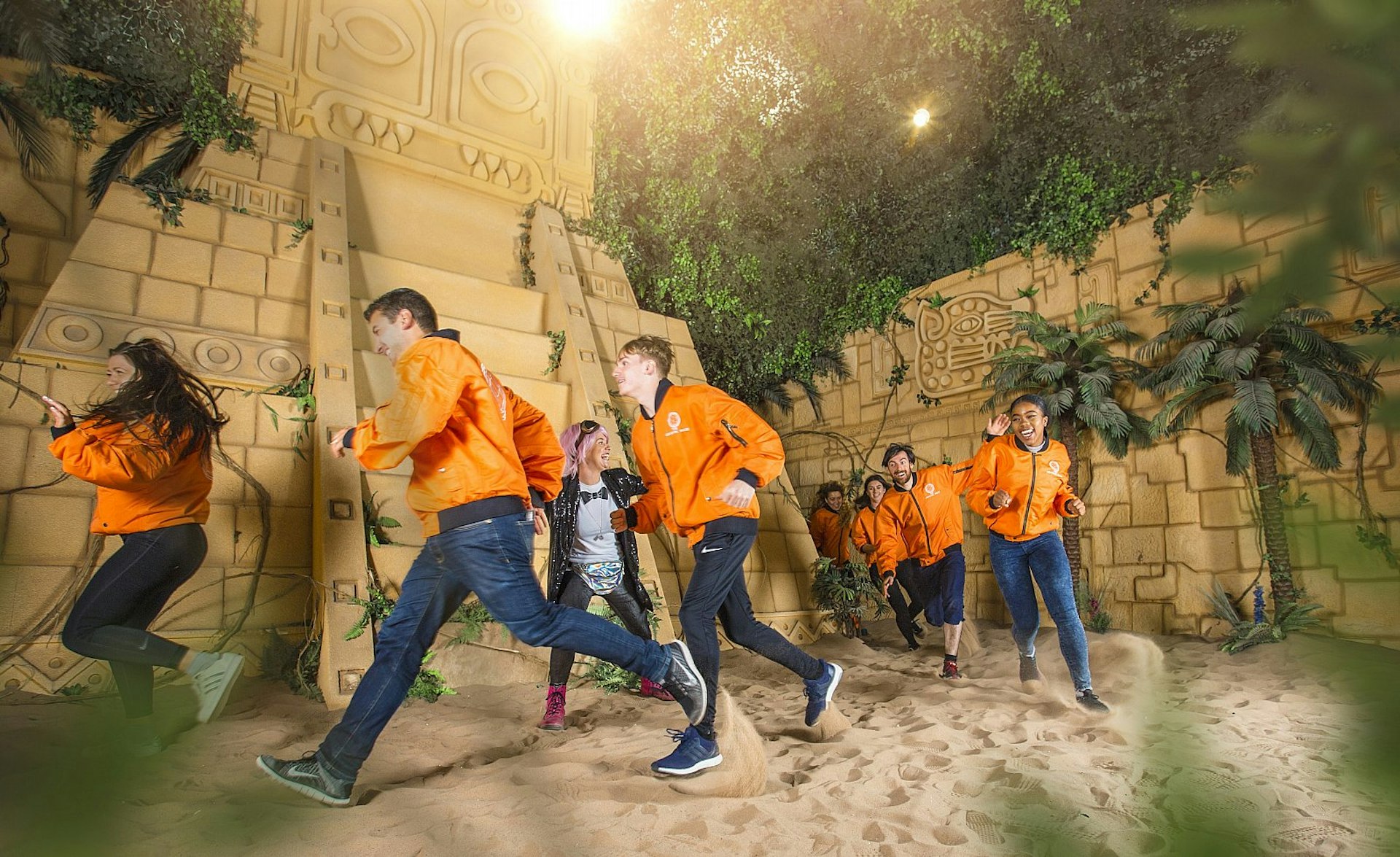 A group of people wearing orange bomber jackets run through a mock-up of an Aztec scene, with sand, a yellow pyramid and walls covered in creepers.