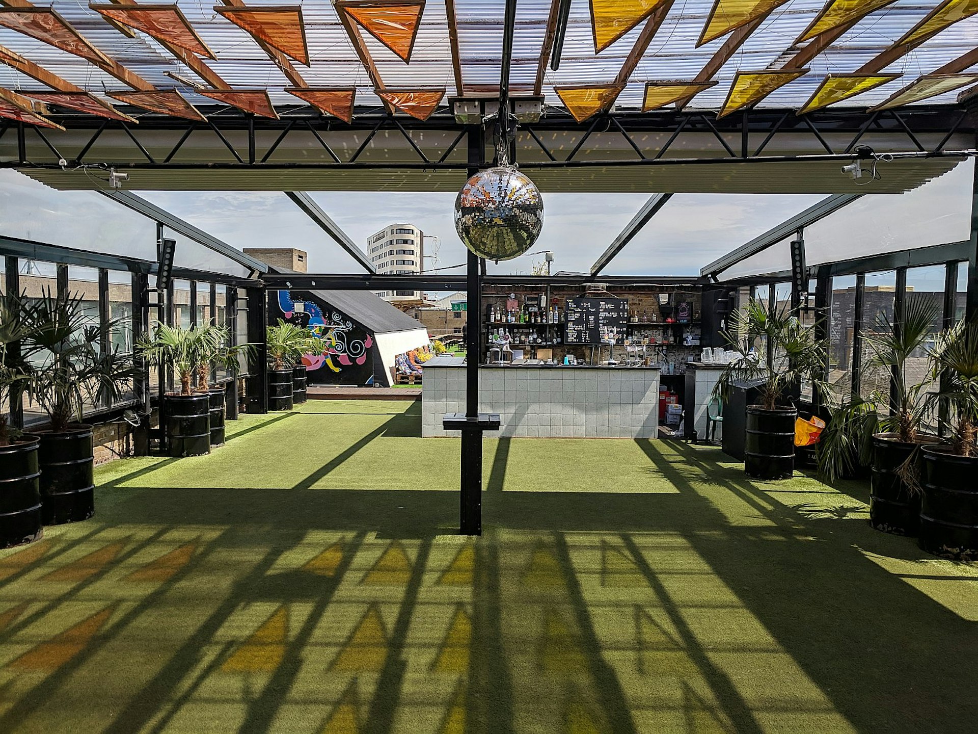An empty Dalston Roof Park; a green floor is flanked by potted plants, while overhead is a glass roof studded with triangular orange and yellow mosaics; a white-tiled bar is in the corner.