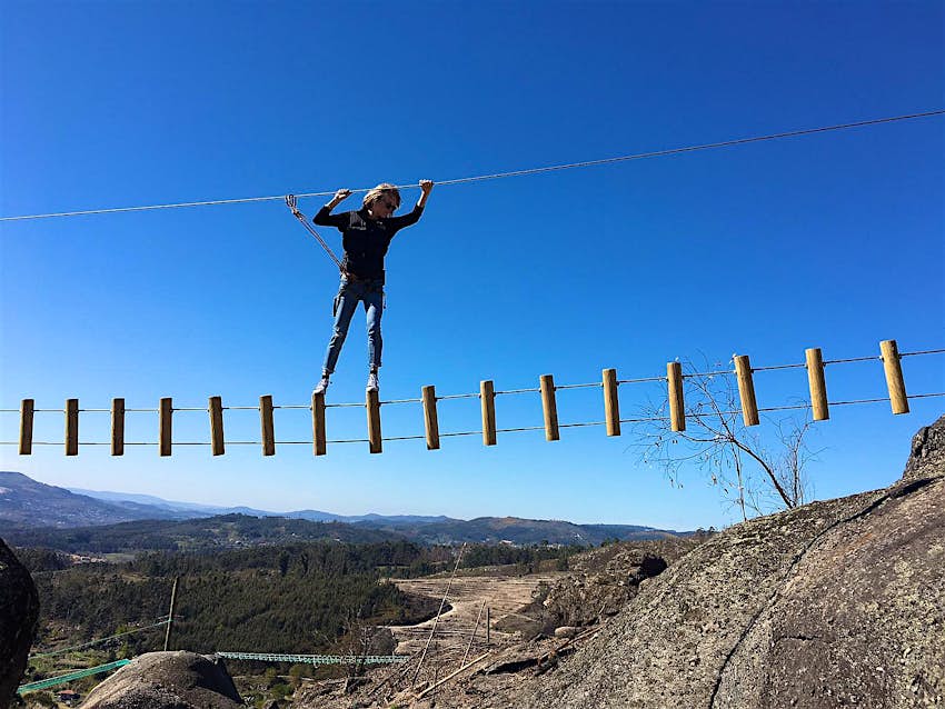 A woman traverses a rope suspension bridge amid rocky hillside scenery.