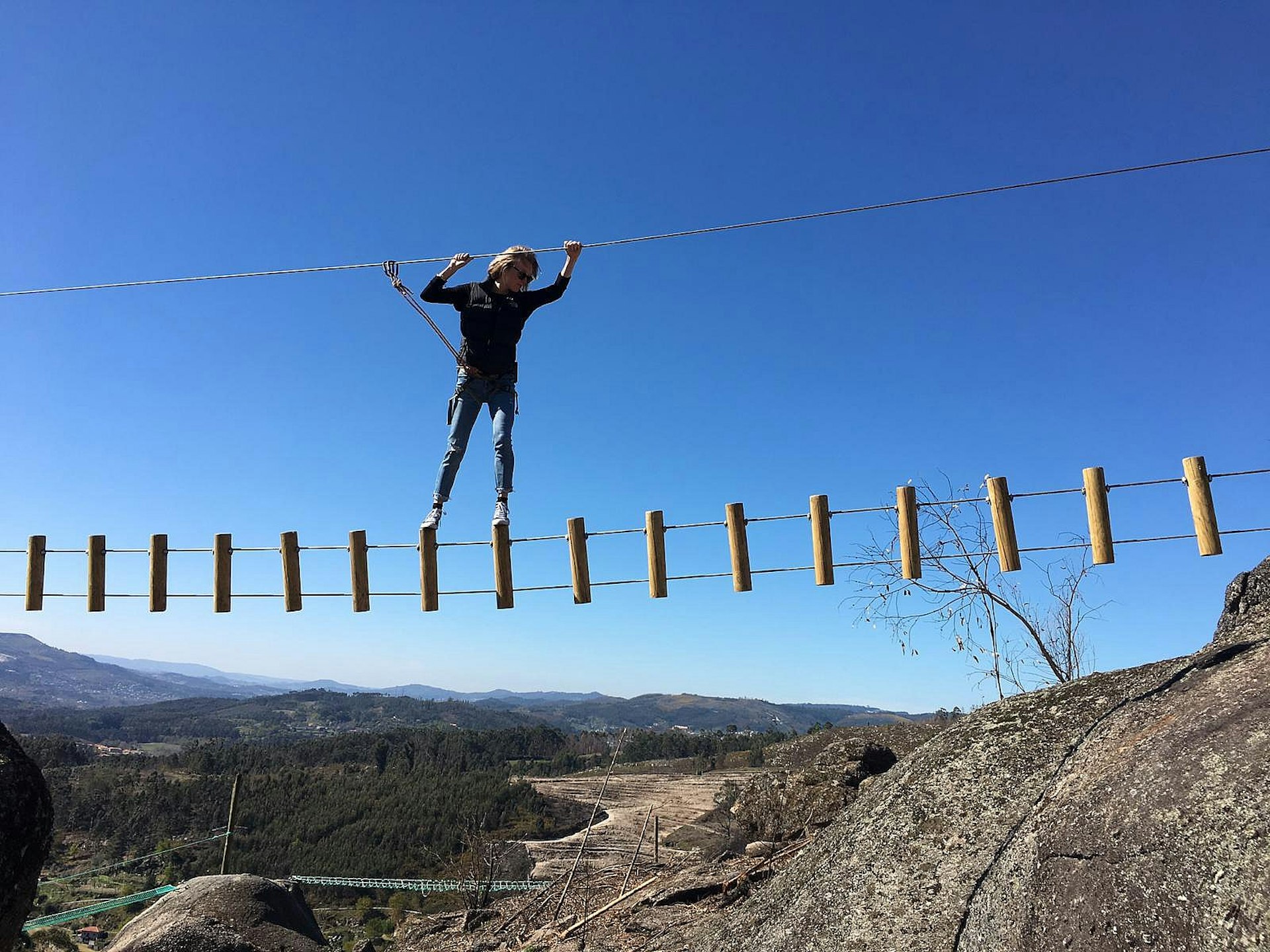A woman traverses a rope suspension bridge amid rocky hillside scenery.
