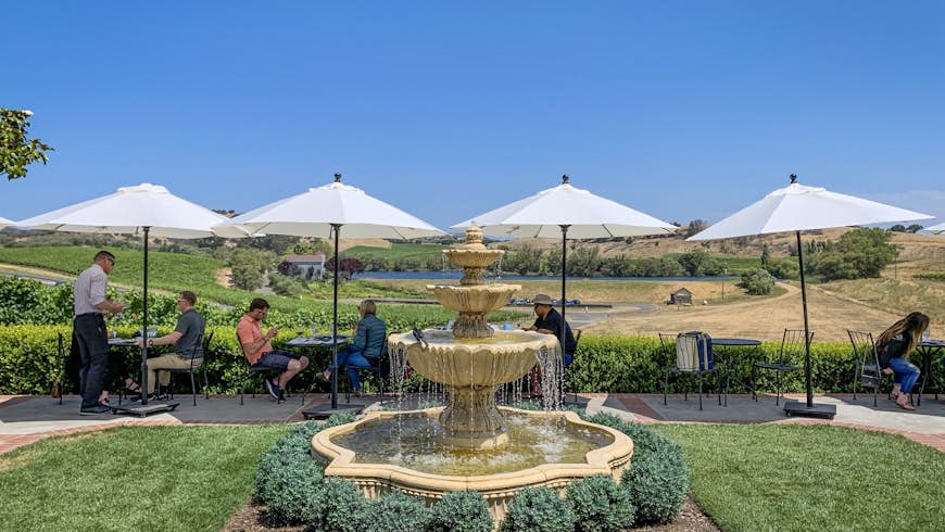 A collection of people sit at tables under umbrellas near a fountain; weekend in Napap