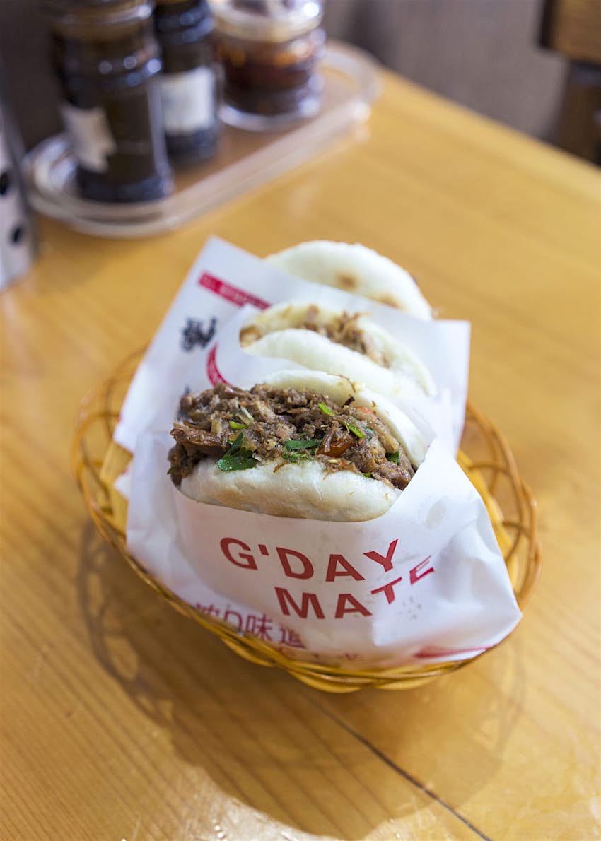 Features - EJ-Fine-Food-eat-curiously A close up shot of rou jia mo served in EJ Fine Food restaurant in Melbourne. It is a type of bao bun, served wrapped in paper that has 'G'DAY MATE' and some script writing in red printed on it, in an orange basket. The white buns are stuffed with brown meat and vegetables.