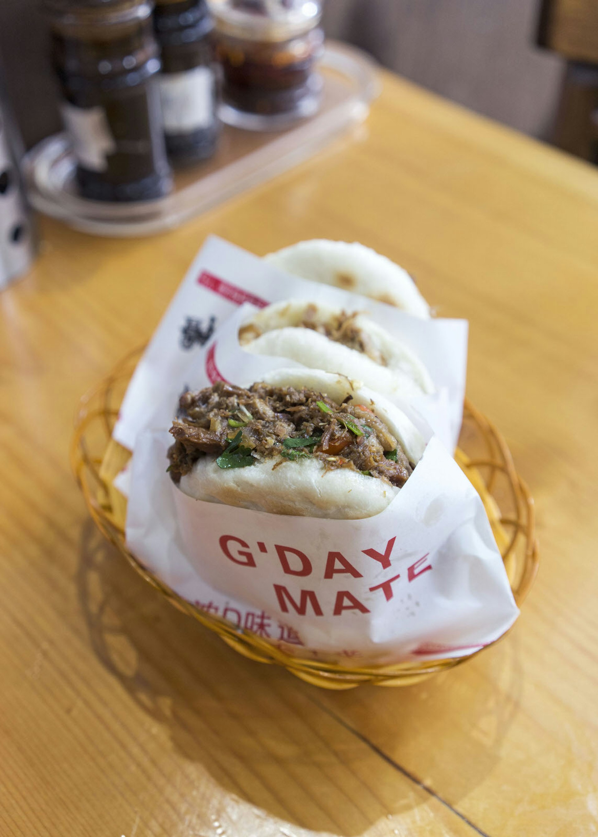 A close up shot of rou jia mo served in EJ Fine Food restaurant in Melbourne. It is a type of bao bun, served wrapped in paper that has 'G'DAY MATE' and some script writing in red printed on it, in an orange basket. The white buns are stuffed with brown meat and vegetables.