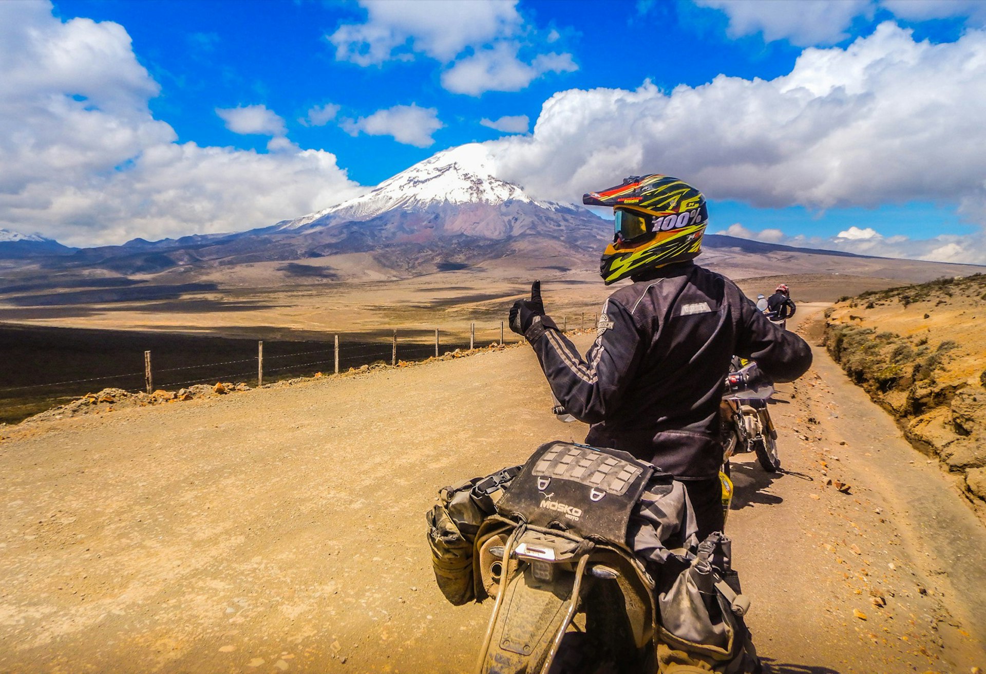a person gives a thumbs up while on a motorcycle with a towering snow capped peak in the back ground on an epic adventure 