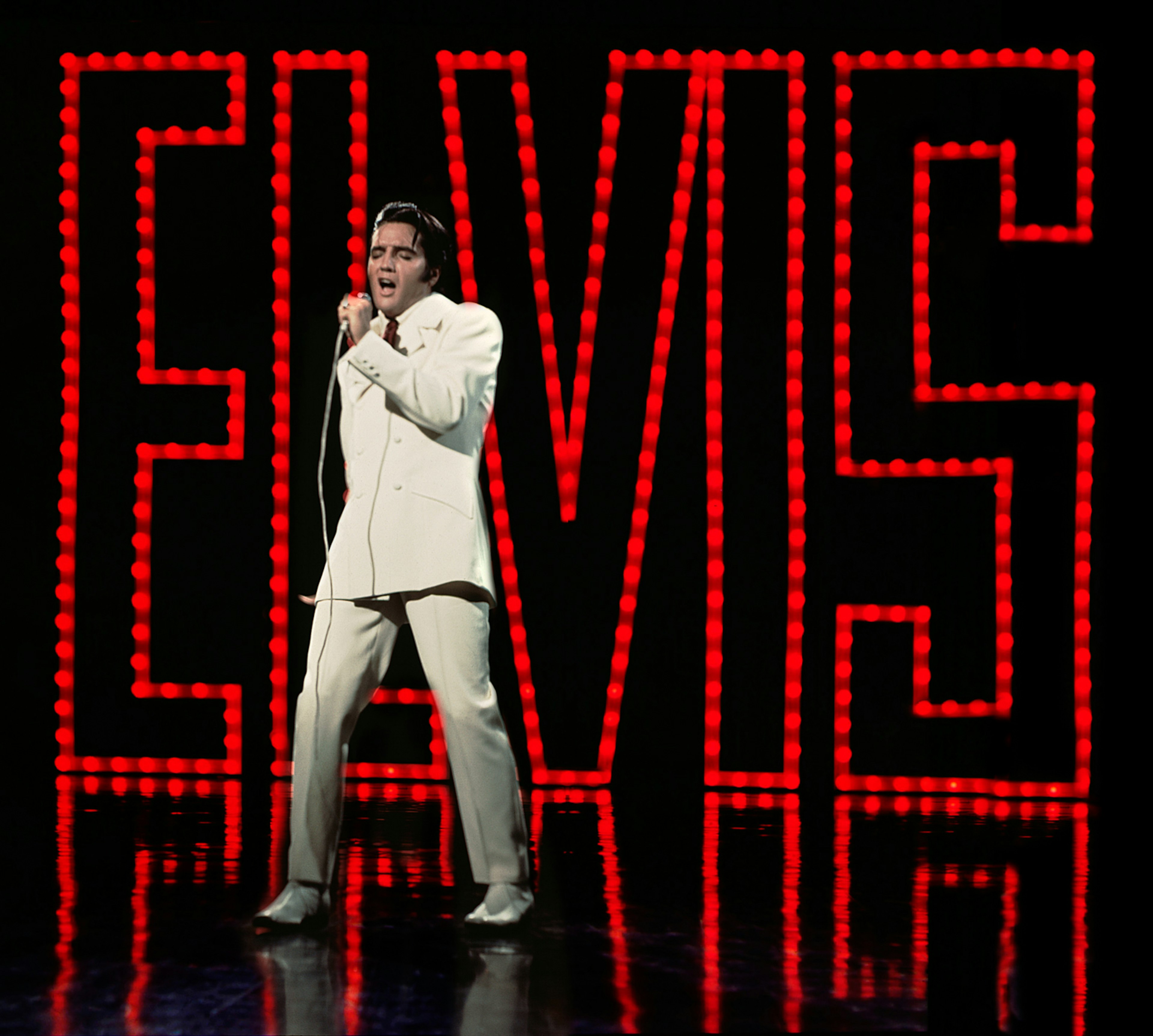 Elvlis Presley in a white suit on stage, in front of a series of red lights that spell ELVIS