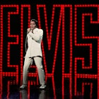 Elvlis Presley in a white suit on stage, in front of a series of red lights that spell ELVIS