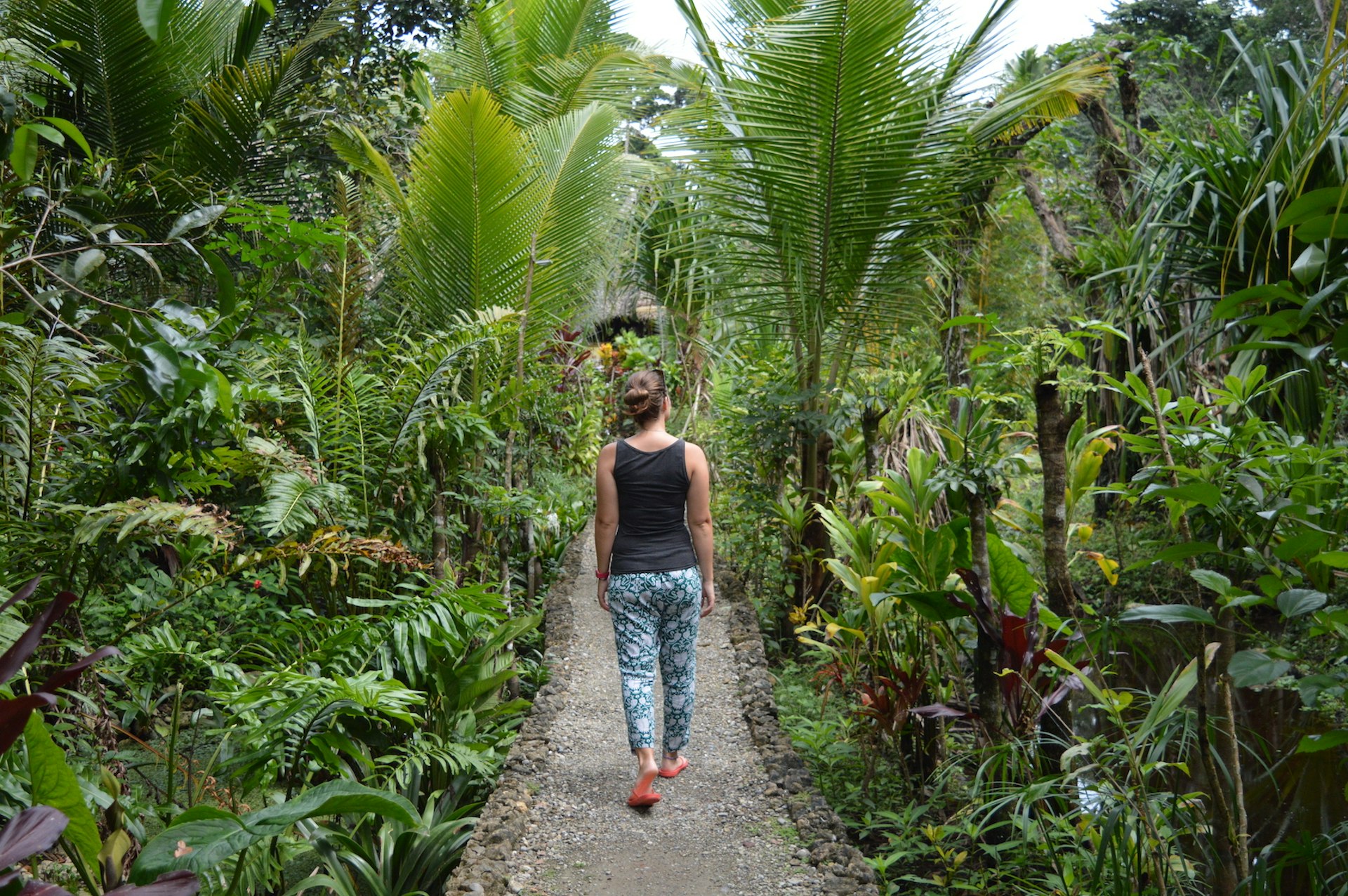 Photo of a woman from behind wearing flip flops, trousers and a tank top as she walks on a dusty path through lush tropical gardens in Guatemala