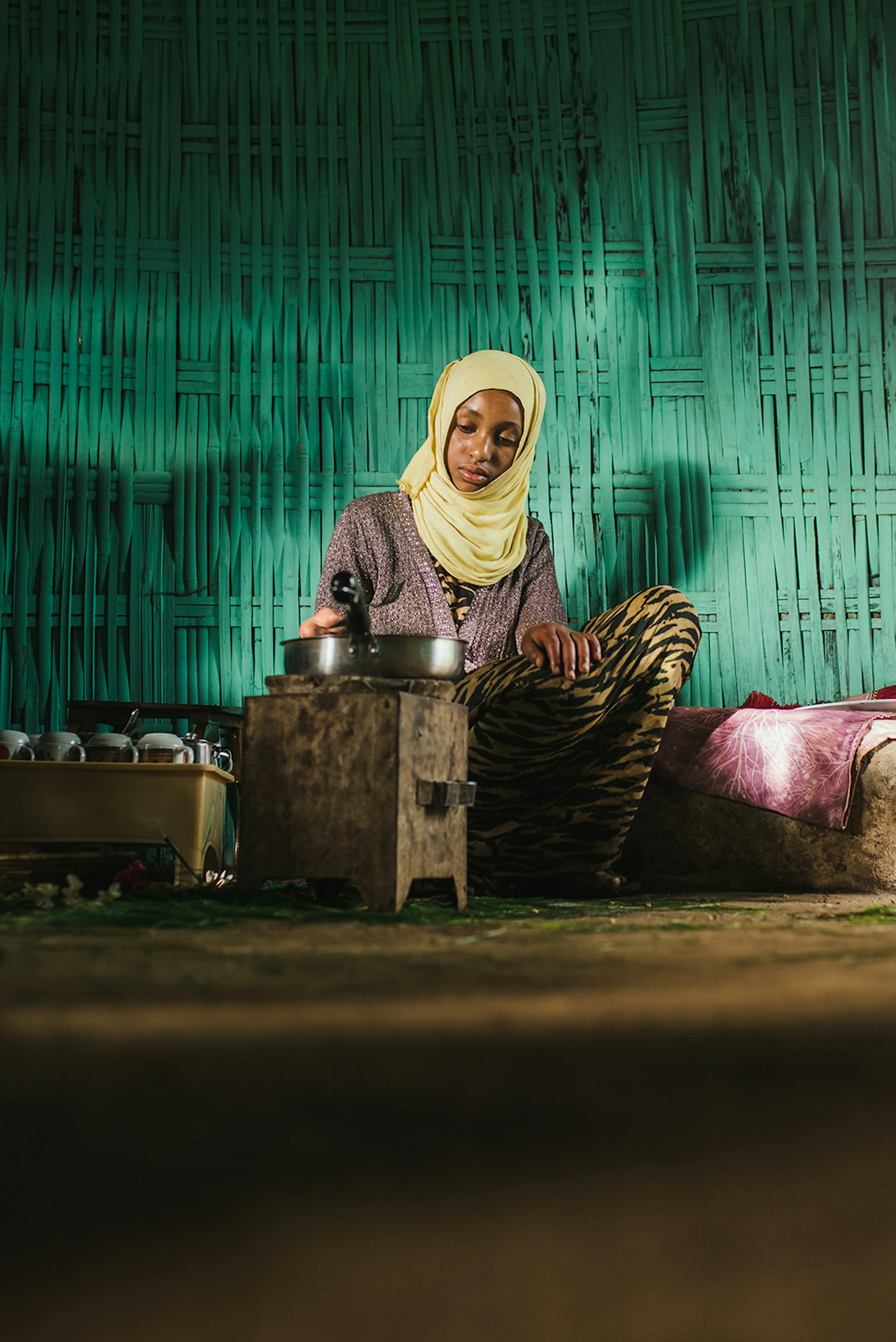 An Ethiopian girl prepares a coffee ceremony; she is wearing a yellow headscarf and sitting in front of a turquoise wall.
