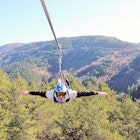A man on a zip line at Pena Aventura, far above treetops with hills beyond; he is facing the camera with arms outstretched.