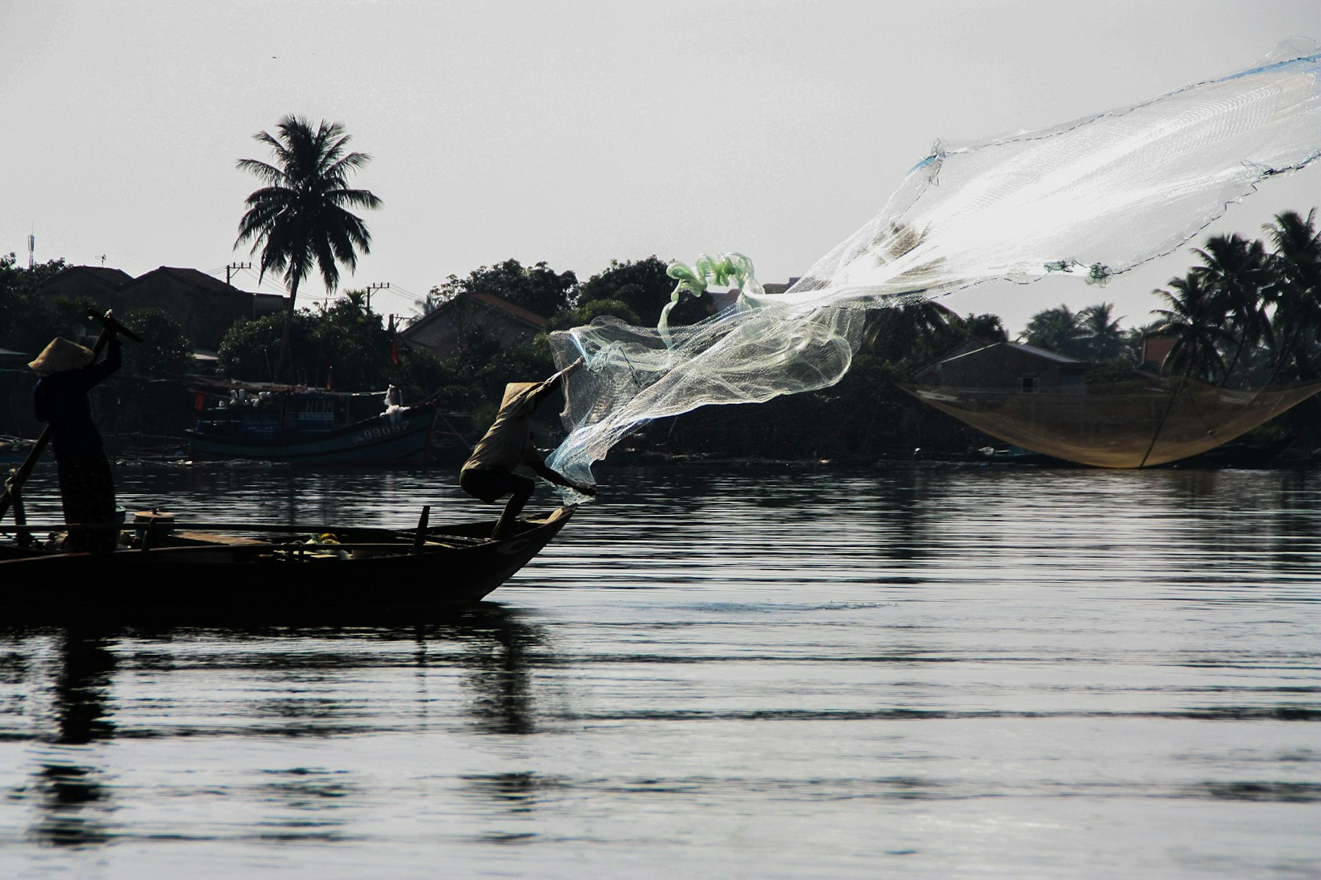 A fisher throws a large fishing net from the end of a small wooden boat in the middle of the river. There are palm trees silhouetted against the grey sky in the background.