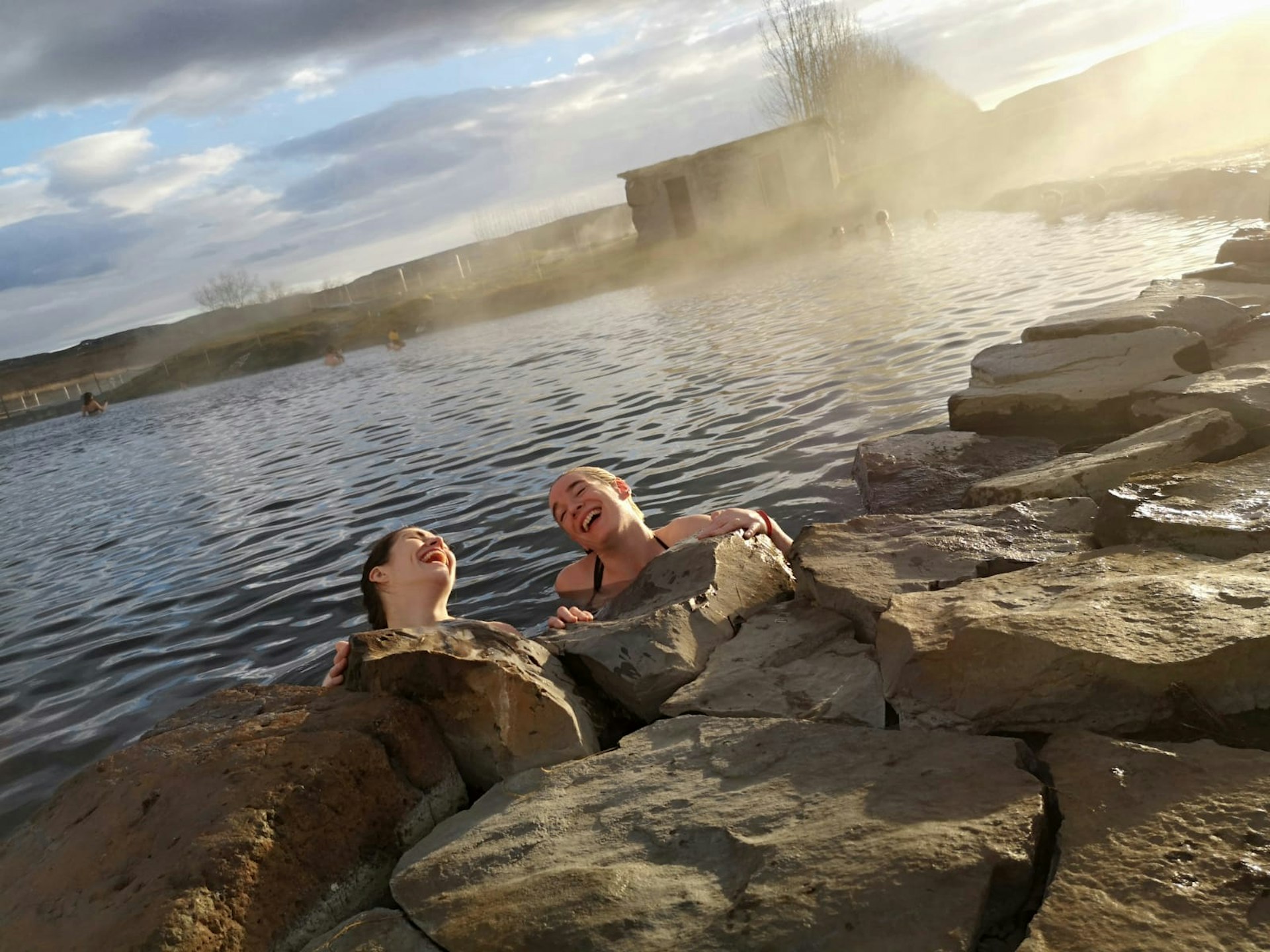 Two women are in a lagoon, resting against some rocks on the side. They both have their heads thrown back in laughter.