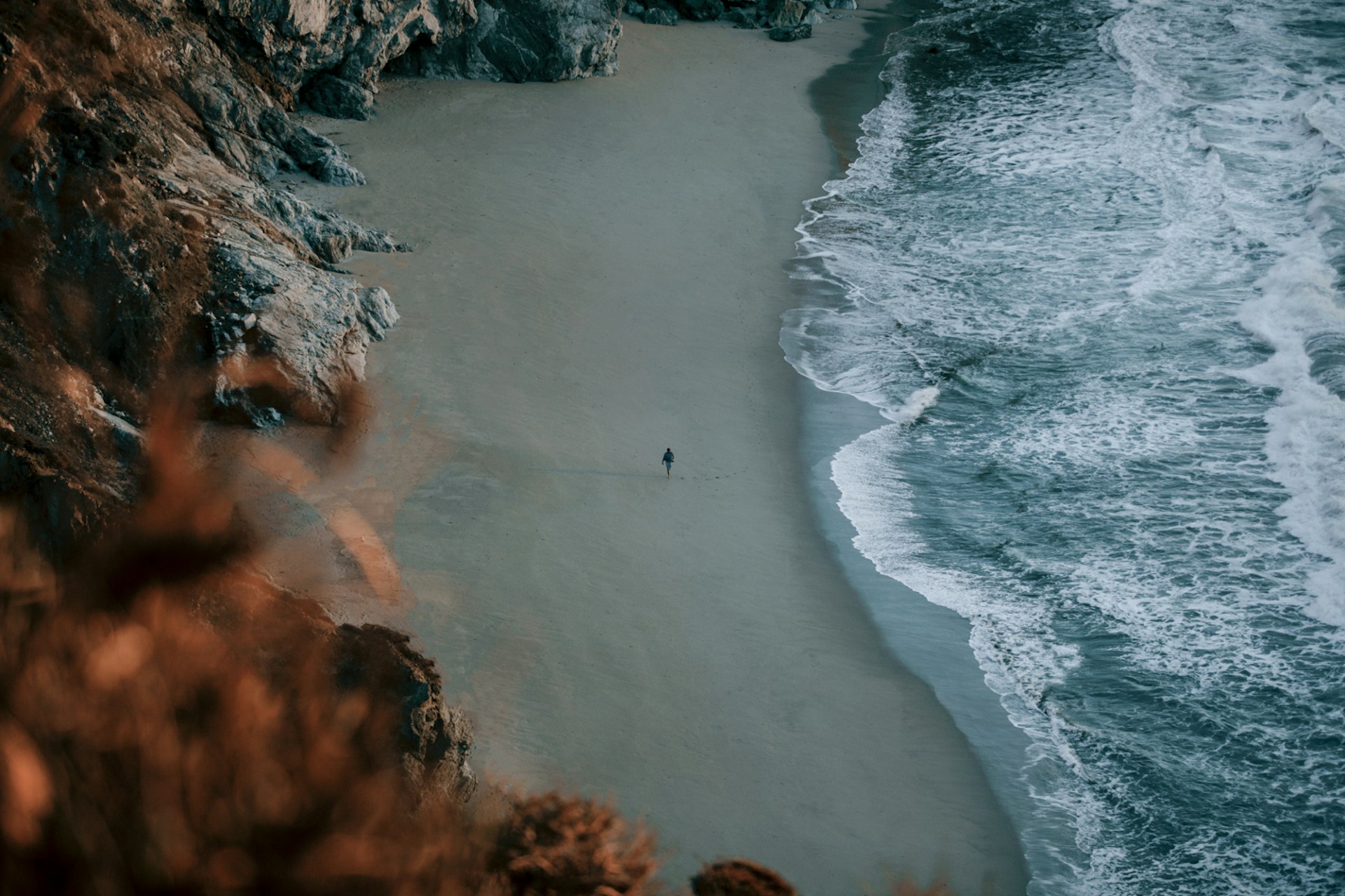 An aerial view of a lone figure walking the beach in Big Sur in California, under a moody light; the sea looks powerful and there are cliffs behind the beach. 