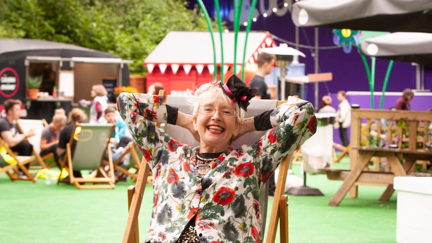English actress and singer Su Pollard poses during a photocall to promote her show 'Harpy' at the Edinburgh Festival Fringe at Underbelly George Square. She is sitting in a deckchair with her hands behind her head and feet raised, there are plastic flowers and the Underbelly venue in the background. 