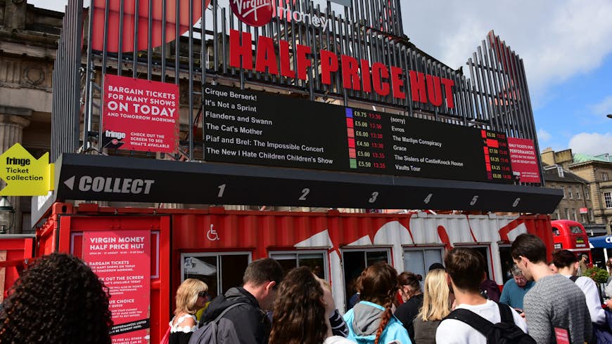 Festival-goers on The Mound precinct queue outside the "Half Price Hut" ticket office during the Edinburgh Festival Fringe. The office is in a shipping container painted red and white with windows to serve customers through. 