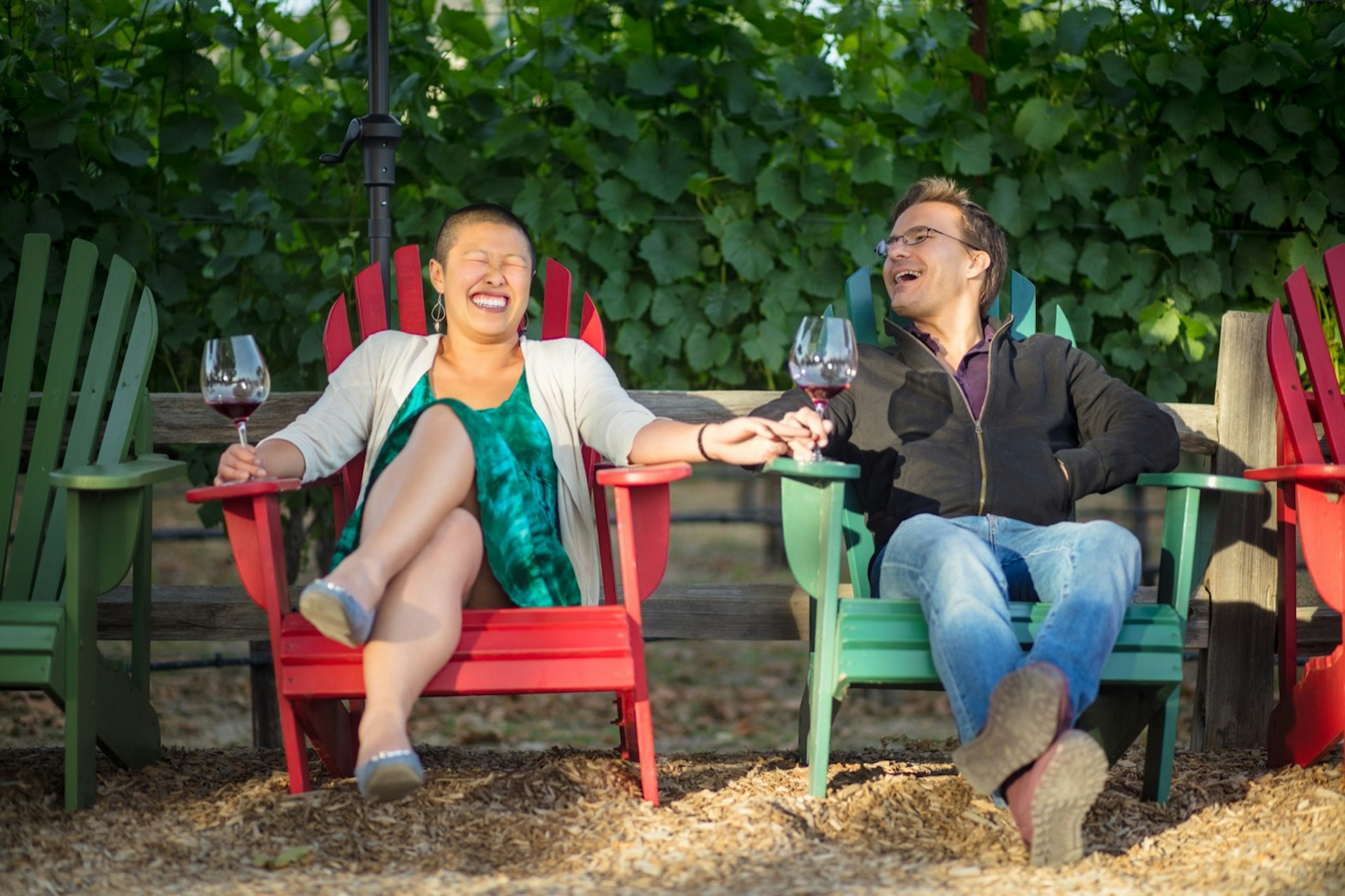 A man and a woman lean comfortably in Adirondack chairs laughing with glasses of wine