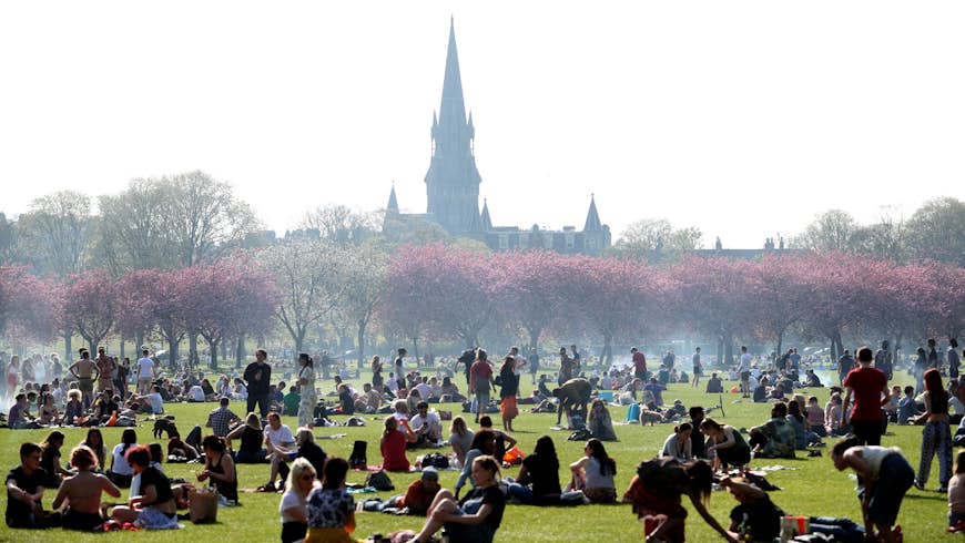 People enjoy the sunshine in The Meadows, Edinburgh. There is smoke from barbecues dotted around the park, there are cherry blossom trees blooming in the background and lots of people sitting on the grass in groups. A church steeple is visible in the background. 