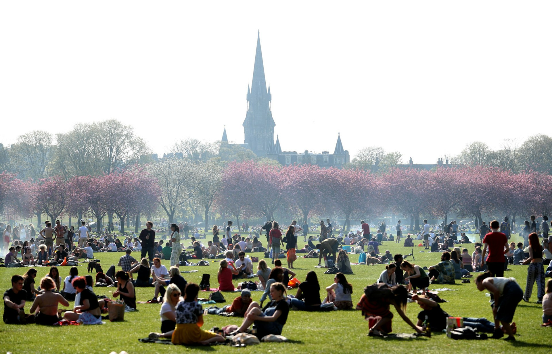 People enjoy the sunshine in The Meadows, Edinburgh. There is smoke from barbecues dotted around the park, there are cherry blossom trees blooming in the background and lots of people sitting on the grass in groups. A church steeple is visible in the background. 