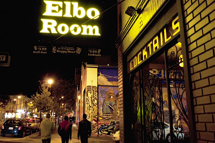 A sign reads Elbo Room over a street where two people walk. Weekend in San Francisco