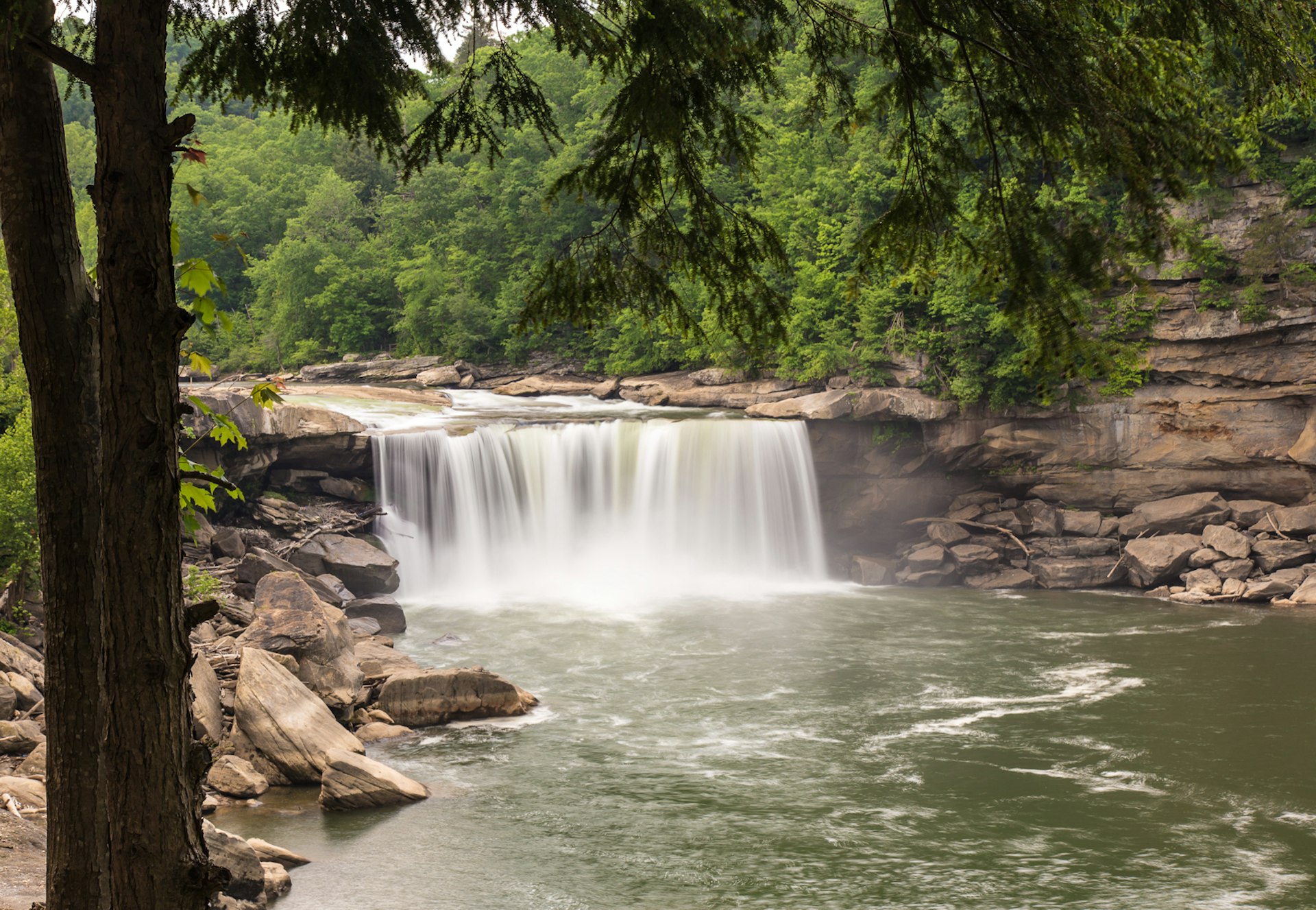 A long exposure of a waterfall, with rocks on either side of the river and green trees in the background; summer trips in the Southern US