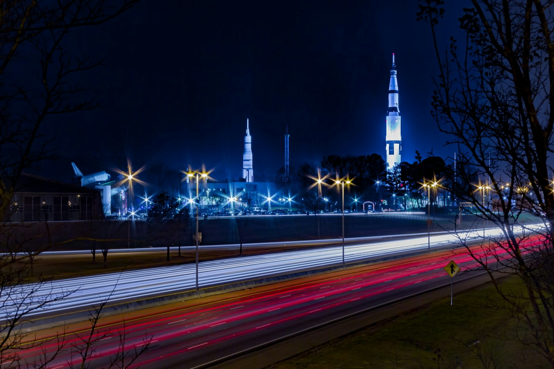 A long exposure shot of US Space and Rocket Center Huntsville, AL with highway traffic. There are parallel streaks of red and white from the car lights in the foreground and the white, needle-like shuttles are visible in the background; Apollo anniversary experiences