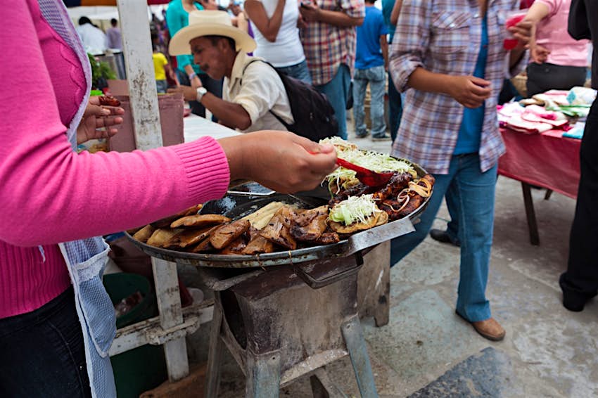 A woman ladles mole sauce over a plate of enchiladas from a street stall while lots of people walk past