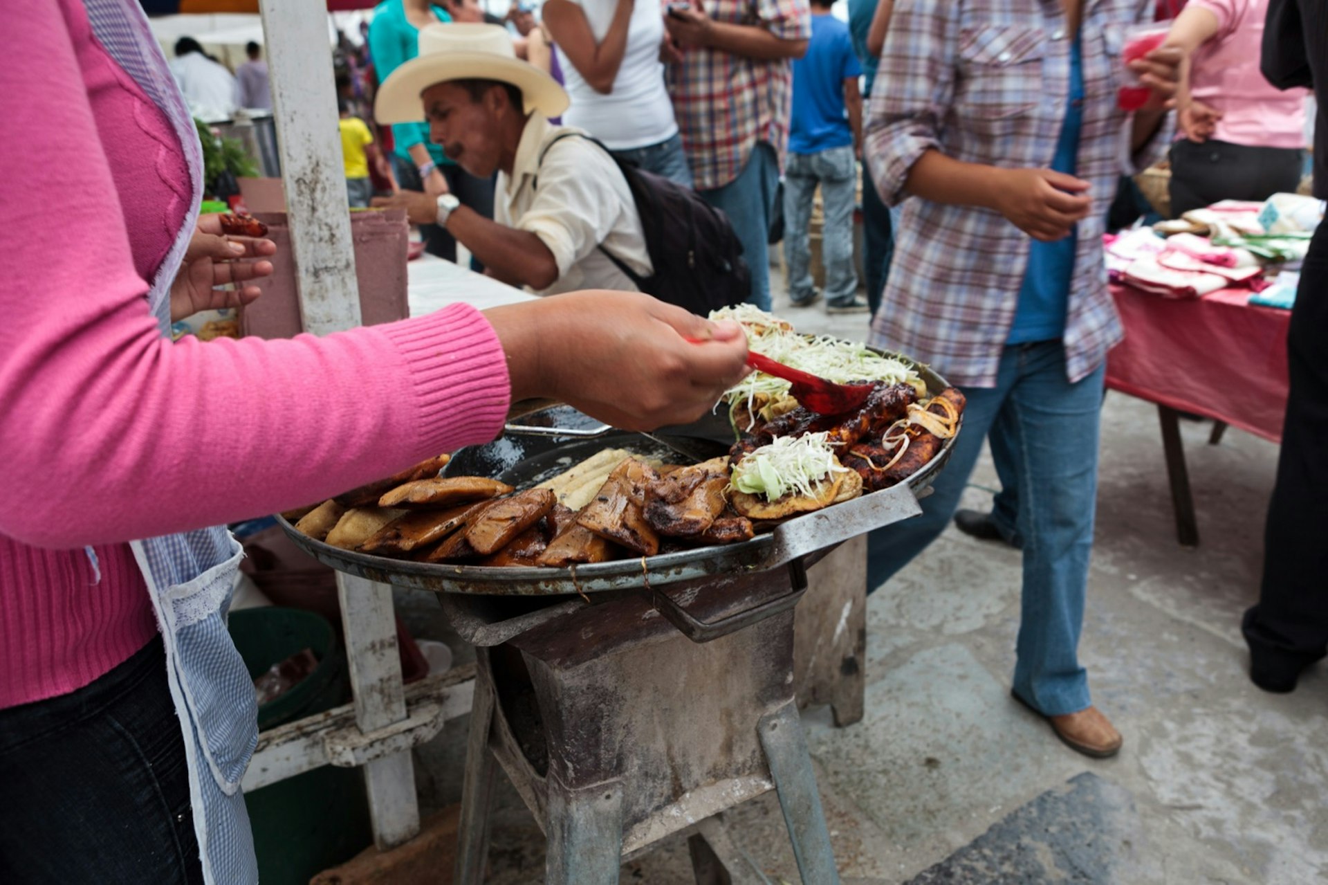 A woman ladles mole sauce over a plate of enchiladas from a street stall while lots of people walk past