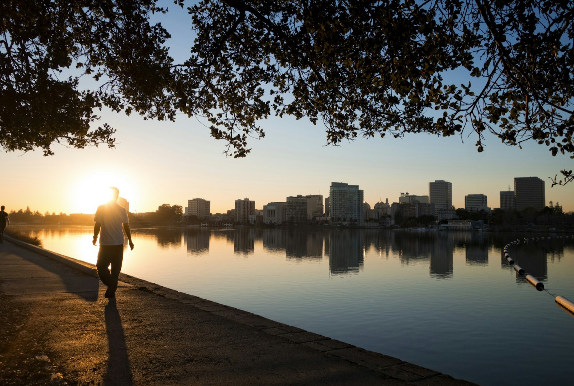 The sun is setting over a lake behind the Oakland skyline which is reflected in the lake a man is walking around