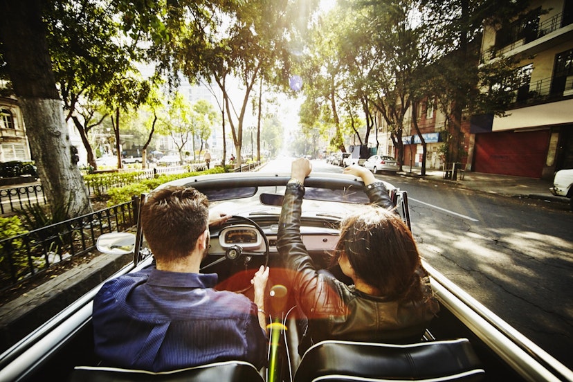 A man drives a convertible car while a woman rides in the passenger seat down a street lined with Jacaranda trees on their way out of town for a day trip from Mexico City