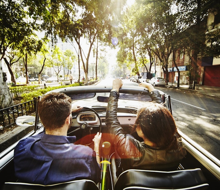 A man drives a convertible car while a woman rides in the passenger seat down a street lined with Jacaranda trees on their way out of town for a day trip from Mexico City
