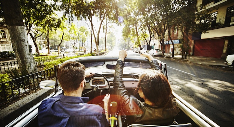 A man drives a convertible car while a woman rides in the passenger seat down a street lined with Jacaranda trees on their way out of town for a day trip from Mexico City