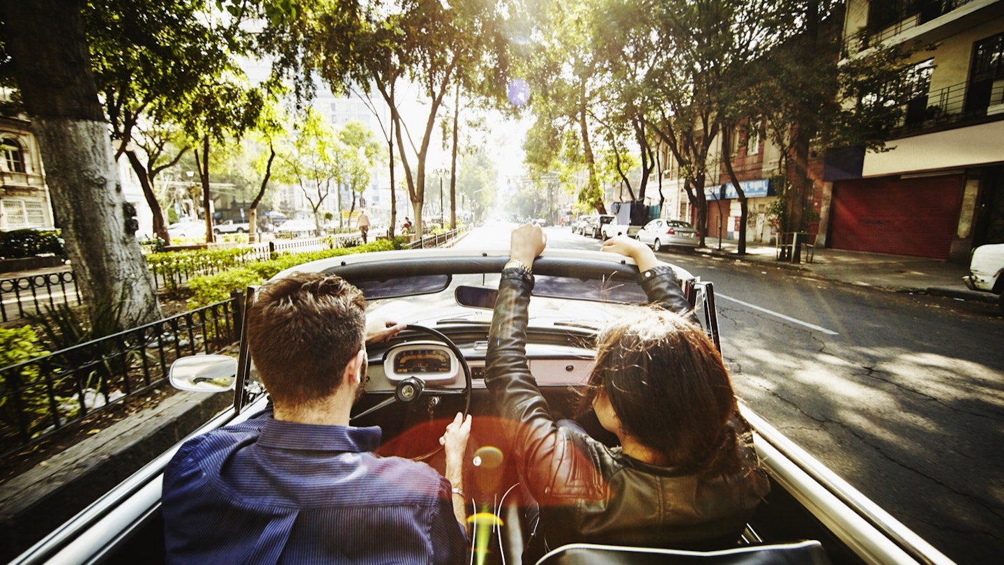 A man drives a convertible car while a woman rides in the passenger seat down a street lined with Jacaranda trees on their way out of town for a day trip from Mexico City